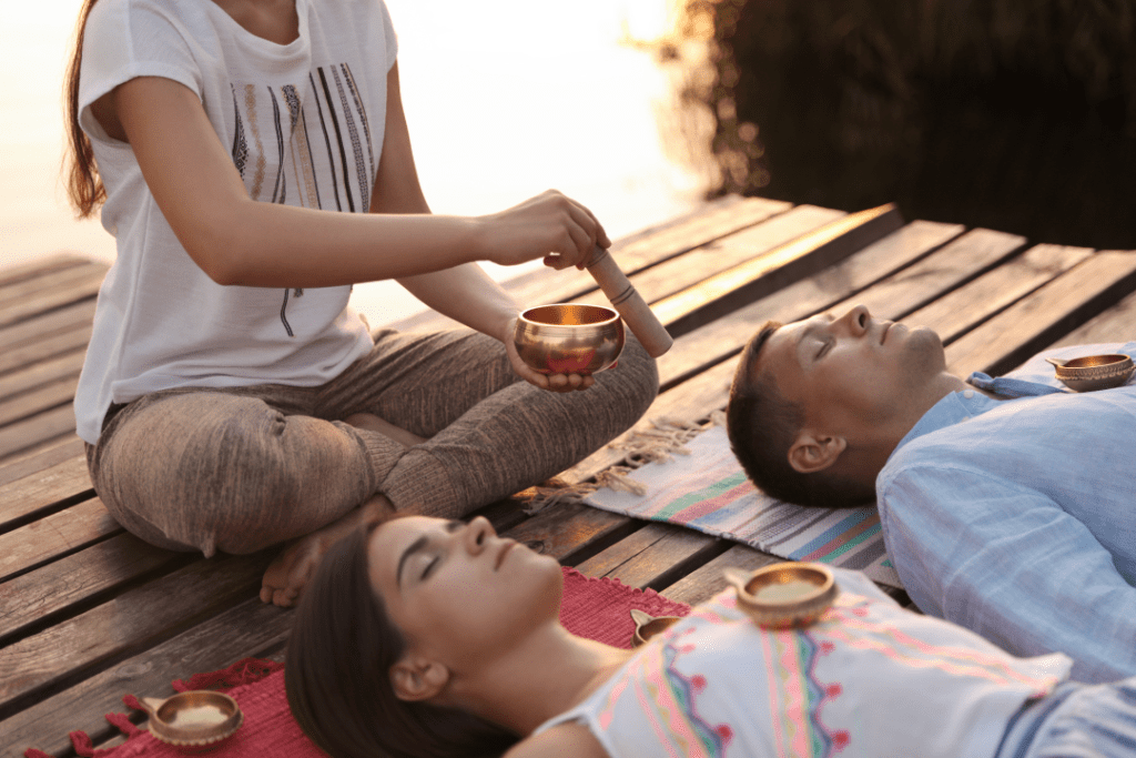 one person is sitting with a tibetan singing bowl and giving treatment to a male and female laying on the ground each with a bowl on their chests