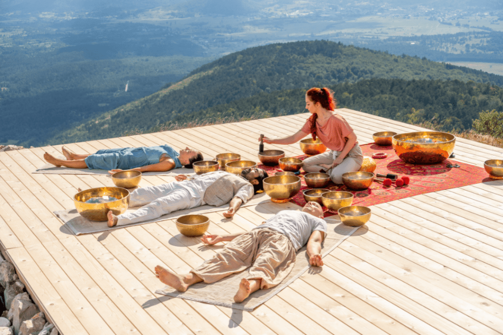 Mountain view in the back with space to work. 3 people laying on a wooden floor getting sound bowl treatment therapy from a female therapist