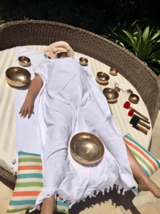 Person laying on her back with a cloth over her face and sound bowls around her.